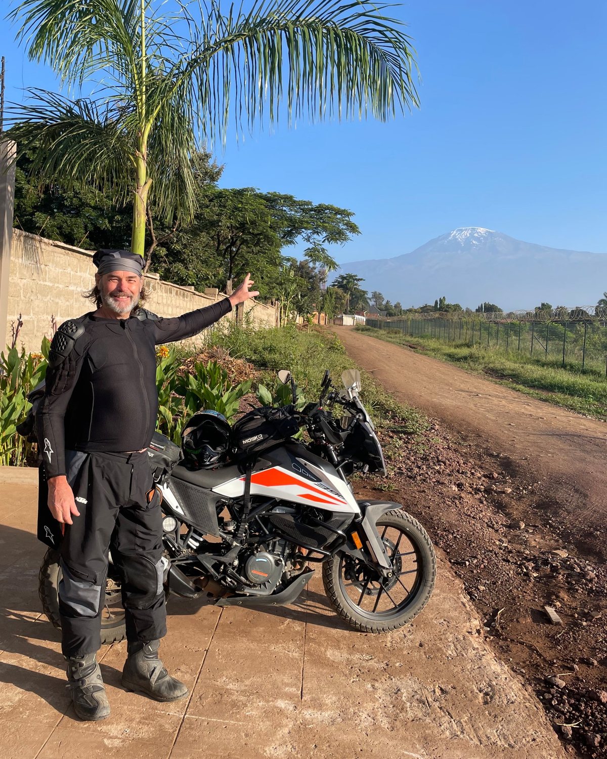 The founder of Tailored Tracks Africa standing by his motorbike on a red-dirt road, with Kilimanjaro behind him
